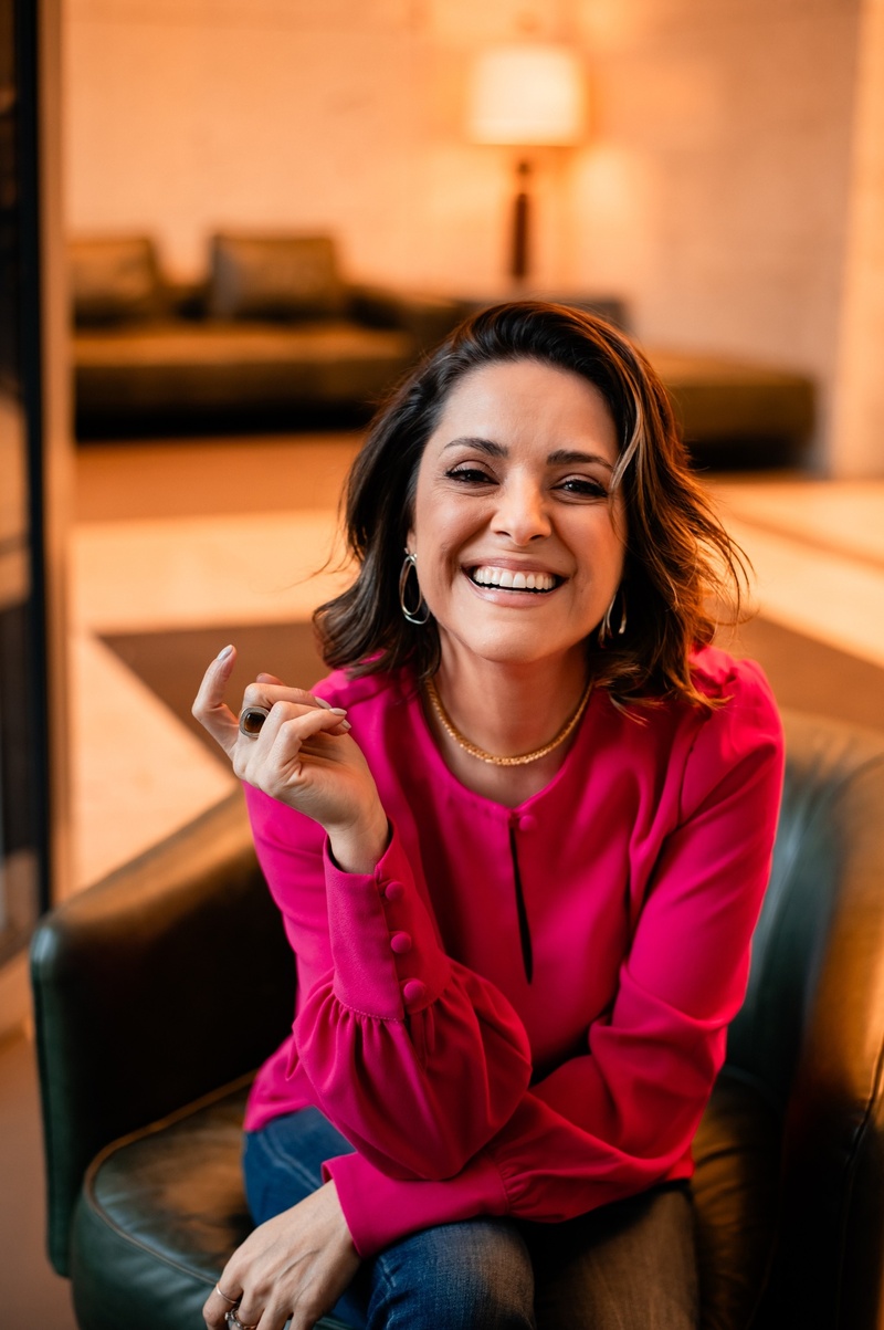 Jana Barcellos sorrindo, sentada em uma poltrona de couro, vestindo blusa rosa.