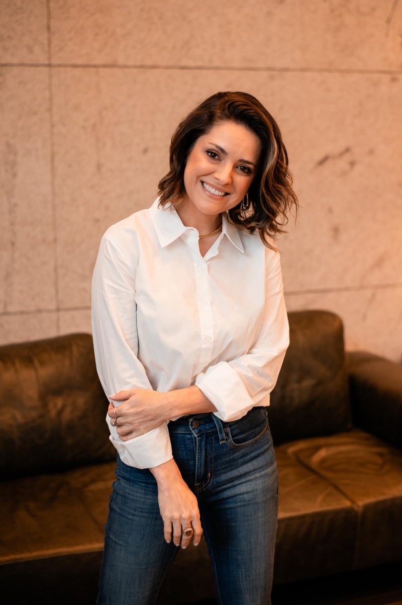 Jana Barcellos em pé, de camisa branca, sorrindo diante de uma parede de pedra.
